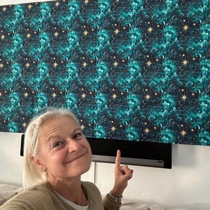 May include: A woman smiles and points to a black soundbar mounted on a white wall. The wall is decorated with a teal and gold patterned fabric with a repeating design of stars and swirls.