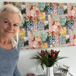 Op de afbeelding: Een vrouw met blond haar glimlacht naar de camera. Achter haar is een muur met een kleurrijk wandkleed met een patchwork van tegels met bloemmotieven. Het wandkleed is ingelijst en hangt aan een witte muur.
