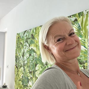 Puede incluir: Una mujer con el pelo corto y rubio sonríe a la cámara. Lleva una camiseta gris claro y un collar. El fondo es una pared con un gran estampado tropical.