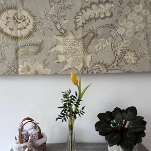 May include: A still-life arrangement featuring a yellow tulip in a clear glass vase, a small potted plant, and a decorative basket with bottles. A floral patterned fabric hangs in the background. The scene is set on a gray surface.