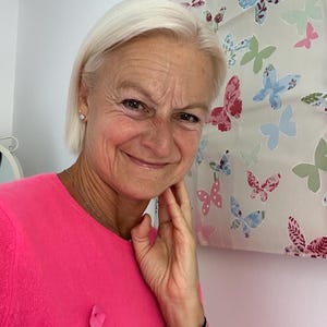 May include: A woman with short, blonde hair is wearing a pink sweater with a pink ribbon pinned to it. She is smiling and has her hand on her chin. She is wearing several bracelets on her wrist.