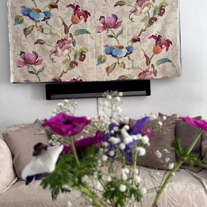 Könnte beinhalten: Ein beigefarbener Stoff mit einem floralen Muster mit rosa, roten, blauen und grünen Blumen. Der Stoff hängt an einer Wand über einer schwarzen Soundbar.