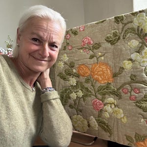 Peut inclure: Une femme aux cheveux blancs courts porte un pull marron clair. Elle se tient devant un tissu beige à motif floral. Le tissu présente des fleurs orange, roses et blanches avec des feuilles vertes.