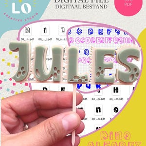 Op de afbeelding: Een gepersonaliseerde taarttopper met de naam JULES in een gelaagd dinosaurus-lettertype. De letters zijn lichtgroen met een lichtroze achtergrond en het ontwerp bevat een puntige prikker. De tekst JULO CREATIVE STUDIO is zichtbaar.