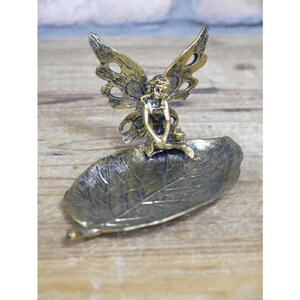 May include: Gold metal fairy figurine sitting on a leaf-shaped dish. The fairy has wings and is holding a flower.