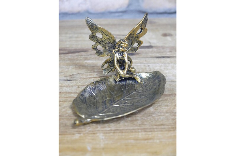 May include: Gold metal fairy figurine sitting on a leaf-shaped dish. The fairy has wings and is holding a flower.