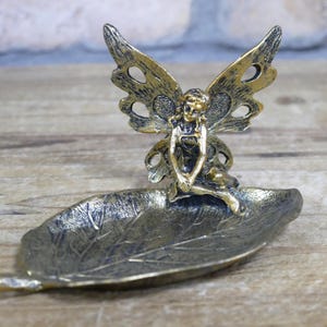 May include: A gold-toned metal fairy figurine with wings sits on a leaf-shaped dish. The fairy is sitting with her legs crossed and her hands resting on her lap. The dish is made of metal and has a textured surface.