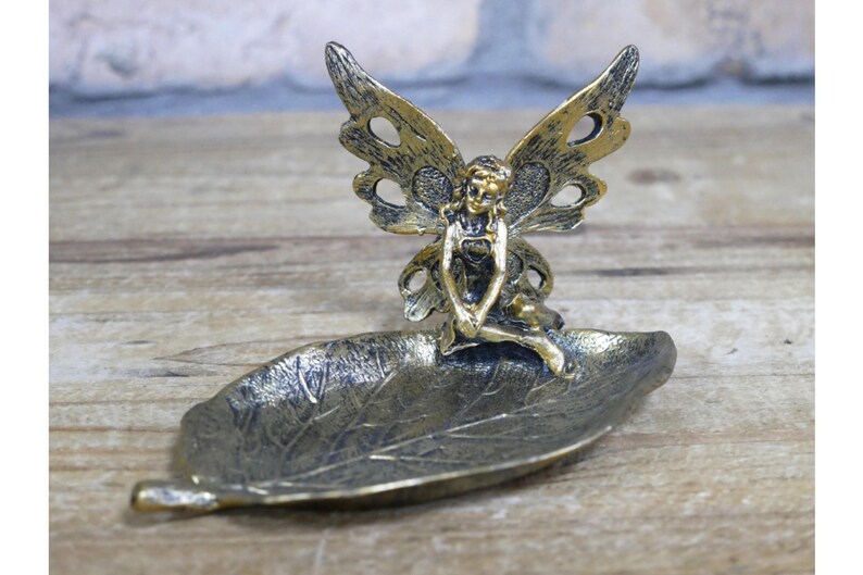 May include: A gold-toned metal fairy figurine with wings sits on a leaf-shaped dish. The fairy is sitting with her legs crossed and her hands resting on her lap. The dish is made of metal and has a textured surface.