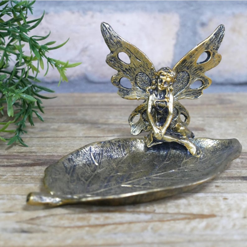 May include: Gold metal fairy figurine with wings sitting on a leaf-shaped dish.