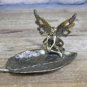 May include: A gold-toned metal trinket dish shaped like a leaf with a fairy figurine sitting on top. The fairy has wings and is holding a flower.