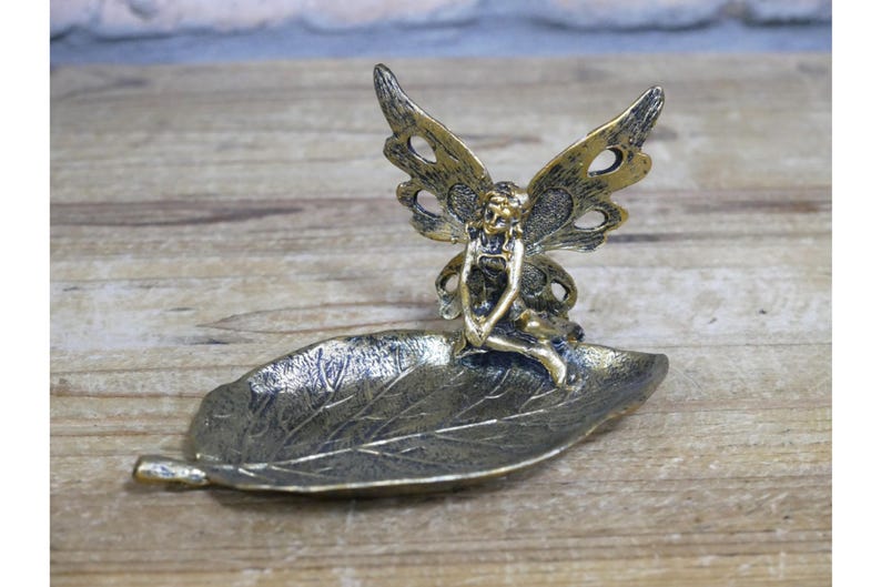 May include: A gold-toned metal trinket dish shaped like a leaf with a fairy figurine sitting on top. The fairy has wings and is holding a flower.