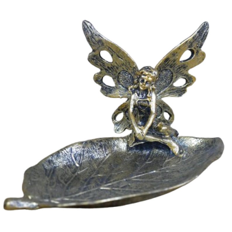 May include: A gold and black metal fairy figurine sitting on a leaf-shaped dish. The fairy has wings and is wearing a dress.