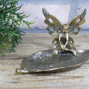 May include: Gold metal fairy figurine with wings sitting on a leaf-shaped dish. The fairy is holding a flower in her hand.