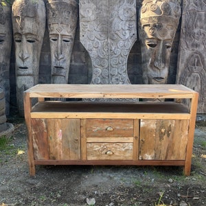 May include: A rustic wooden cabinet with two doors and two drawers. The cabinet is made of reclaimed wood and has a natural finish. The cabinet has a shelf above the drawers and is sitting in front of a wall with carved wooden panels.