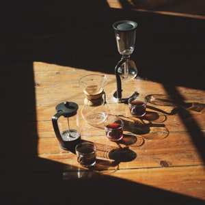 Peut inclure: Une cafetière noire et argentée avec une carafe en verre et une presse à café noire. Il y a quatre petites tasses en verre remplies de café sur une surface en bois.