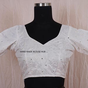 May include: White cotton blouse with short sleeves and a V-neckline. The blouse is decorated with small, round, mirrored embellishments. The text "HAND MADE BLOUSE HUB" is printed on the front of the blouse.