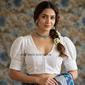 May include: A white, V-neck blouse with short, puffed sleeves. The blouse has a textured pattern and the text "HAND MADE BLOUSE HUB" is visible. The woman is wearing a silver necklace and earrings. Her hair is styled with flowers.