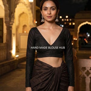 Puede incluir: Una blusa negra de manga larga con cuello en V profundo y el texto "HAND MADE BLOUSE HUB". La blusa se combina con una falda drapeada marrón. La imagen está tomada de noche con elementos arquitectónicos de fondo.