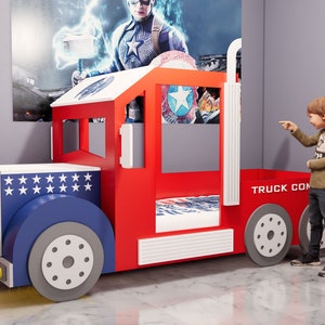 Könnte beinhalten: Ein rotes und blaues Spielzeug-LKW-Bettgestell mit einer weißen Matratze und einer weißen und blauen Bettwäsche. Der LKW hat ein Tankstellenthema mit einer Zapfsäule und einem Schild mit der Aufschrift "Tankstelle". Ein kleiner Junge steht neben dem LKW-Bettgestell und zeigt auf die Zapfsäule.