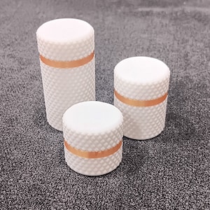 May include: Three white knobs with a gold band around the middle. The knobs are different sizes and are arranged in a row from largest to smallest.