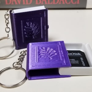May include: Two purple book-shaped keychain holders with a silver chain. The keychain holders open to reveal a small compartment for a memory card. One holder is open and shows a SanDisk memory card inside.