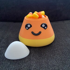 May include: A 3D-printed candy corn-shaped container with a smiling face, filled with candy corn. The container is orange, yellow, and white, with a separate white lid. A fun, festive item for Halloween.