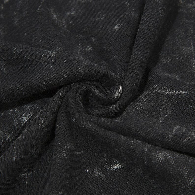 May include: Close-up of a black fabric with a distressed, mottled appearance. The material is tightly woven, with a slightly textured surface and subtle variations in tone, creating a vintage or worn aesthetic. The fabric is gathered, showing folds and shadows.