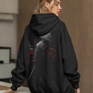 Könnte beinhalten: Schwarzer Kapuzenpullover mit einem Samurai-Krieger-Motiv, der einen Hut und ein Schwert trägt. Das Design enthält japanischen Text und rote florale Akzente. Der Hoodie hat eine lockere Passform.