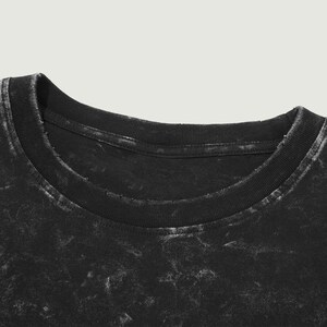 May include: Close-up of a black t-shirt with a crew neck. The fabric has a distressed, faded appearance with white accents, giving it a vintage look. The collar is ribbed and shows signs of wear.