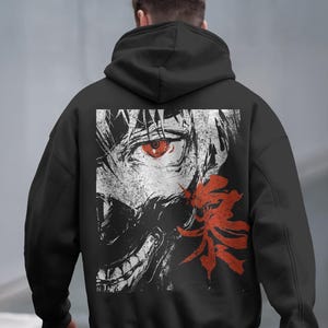 Puede incluir: Sudadera con capucha negra con un diseño gráfico en la espalda. El diseño incluye un ojo rojo, una cara estilizada y un elemento floral rojo. La obra de arte es en blanco y negro con acentos rojos.