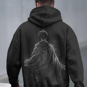 May include: Black hoodie featuring a detailed grayscale graphic on the back. The graphic depicts a figure in a cloak with a symbol. The hoodie has a hood and long sleeves. The overall design is dark and detailed.