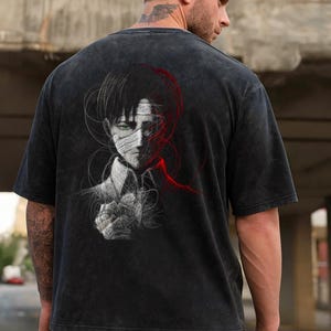 Könnte beinhalten: Dunkelgraues T-Shirt mit einer detaillierten Illustration eines Charakters mit grünen Augen und einem Verband im Gesicht. Das Design enthält rote und weiße Akzente. Das Shirt ist kurzärmlig und für den Freizeitgebrauch.