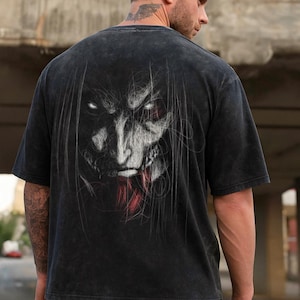 Könnte beinhalten: Schwarzes T-Shirt mit einer detaillierten Monstergesicht-Illustration auf dem Rücken. Das Design verwendet weiße und rote Akzente, die mit dem dunklen Stoff kontrastieren. Das T-Shirt hat ein Vintage-Aussehen. Das T-Shirt ist aus weichem Material.