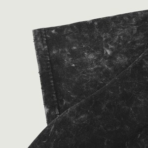 May include: Close-up of a section of black fabric with a distressed, mottled appearance. The fabric has a textured surface with lighter, faded areas, creating a worn, vintage aesthetic. The edge of the fabric is visible, showing a seam.