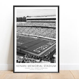 Op de afbeelding: Zwart-wit ingelijste print van Kenan Memorial Stadium in Chapel Hill, North Carolina. Het stadion is gevuld met toeschouwers en het veld heeft de tekst "Tar Heels". De print bevat de naam van het stadion en de coördinaten.
