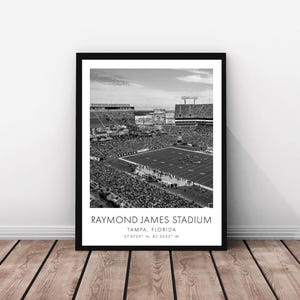 Puede incluir: Fotografía aérea en blanco y negro del Raymond James Stadium en Tampa, Florida. El estadio está lleno de gente y el campo es visible en el centro. El texto "Raymond James Stadium, Tampa, Florida, 27.9759° N, 82.5033° W" está impreso debajo de la imagen.
