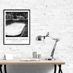 May include: Black and white framed print of the Montreal Forum hockey arena. The print includes the text "MONTREAL FORUM" and coordinates. The image is displayed on a light wood desk with a silver desk lamp.