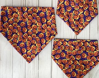 May include: Three Halloween-themed bandanas featuring a repeating pattern of orange jack-o'-lanterns on a purple background with spiderwebs. The bandanas are triangular and made of fabric.
