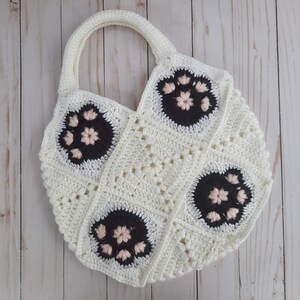 May include: A white crocheted tote bag with black and pink floral accents. The bag has a round bottom and a single handle.