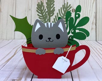 Könnte beinhalten: Eine bezaubernde Papierbastelarbeit mit einer grauen Katze mit weißen Details in einer roten Teetasse mit goldenem Rand. Ein weißes Teebeutel-Etikett hängt an der Tasse. Grüne Papierblätter und Zweige bilden den Hintergrund.