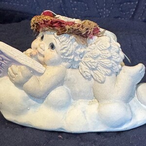 May include: A white cherub figurine with wings, wearing a red and brown wreath, sits on a cloud holding a book that says "Diary".