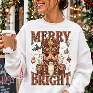 May include: A white crewneck sweatshirt featuring a graphic of a nutcracker wearing a red and green plaid uniform. The text MERRY BRIGHT is printed in red and green above and below the nutcracker, surrounded by ornaments and holly.