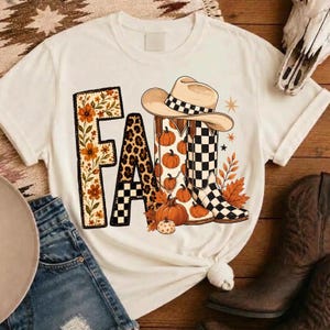 Puede incluir: Una camiseta de manga corta color crema con la palabra FALL en letras grandes. La F está rellena de flores naranjas, la A con estampado de leopardo, la L con un patrón de cuadros y la L con un gráfico de una bota de vaquero y calabazas.