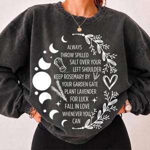 Peut inclure: Un sweat-shirt à col rond gris anthracite avec une impression graphique blanche représentant des phases lunaires, des brins botaniques, un cœur et une salière. Le texte indique : ALWAYS THROW SPILLED SALT OVER YOUR LEFT SHOULDER KEEP ROSEMARY BY YOUR GARDEN GATE PLANT LAVENDER FOR LUCK FALL IN LOVE WHENEVER YOU CAN.