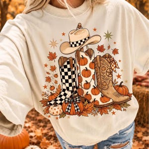 Puede incluir: Una camiseta de color crema con un estampado gráfico de un sombrero de vaquero y tres botas vaqueras. Las botas están decoradas con patrones de calabazas, un diseño de cuadros y motivos florales. El diseño incluye hojas de otoño naranjas.