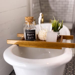 May include: A miniature white clawfoot bathtub with a wooden tray holding a jar of bath salt, a loofah, a candle, and a small plant. The jar has a cork stopper and a label that reads "Bath Salt". The floor has a black and white patterned tile.