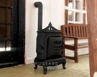 Könnte beinhalten: Schwarzer gusseiserner Holzofen mit einem Rohr und einem dekorativen Oberteil. Der Ofen steht auf einem Ziegelboden vor einer weißen Wand und einer Holztür. Der Text "MUSTARD Miniatures" befindet sich auf der Vorderseite des Ofens.