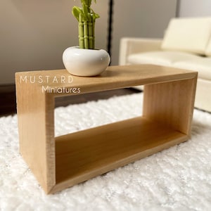 May include: A miniature wooden coffee table with a light brown finish. A small white ceramic pot with a green bamboo plant sits on top. The text "MUSTARD Miniatures" is visible on the table.