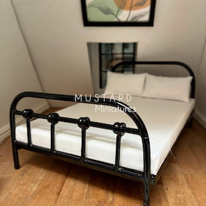 May include: A miniature black metal bed frame with a glossy finish, featuring a rounded headboard and footboard with vertical bars. The bed is dressed with a plain white mattress and two matching white rectangular pillows. Text reads MUSTARD Miniatures.
