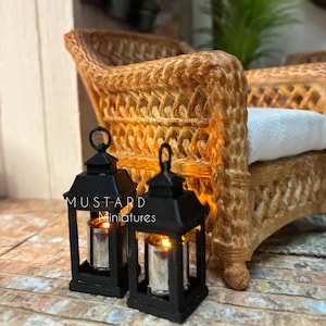 May include: Two miniature black lanterns with glowing orange lights inside, positioned in front of a brown woven wicker armchair with a white seat cushion. The text MUSTARD Miniatures is printed in white on the front of the lanterns.